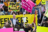  A white-haired white man with glasses and wearing a blazer is speaking and holding a microphone. Around him is a crowd of people holding cameras, protest signs and a banner.