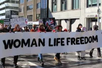 Six marchers carry a long white banner with black capital letters that read WOMAN LIFE FREEDOM down a Toronto city street. Behind them, a parade of women carry other protest signs for international Woman's Day.