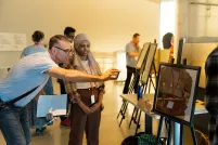 Students in a museum gallery display their work on easels. The focus is on one student talking with an adult who is smiling and taking a photograph with their phone of the student’s work. The artwork shows four people in a hallway with four doors, each with a sign on it.