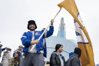 A smiling man in a crowd of people holding a large yellow flag with a blue and white, “W” in the centre. The man is wearing a blue Winnipeg Blue Bombers jacket as he stands in front of the Canadian Museum for Human Rights.