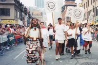 A group of marchers carrying signs that read “2-Spirited People of the 1st Nations” are walking down the centre of a city street while onlookers watch behind barriers on both sides.