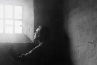 A man stands in dark shadows in a stark stone cell, gazing upward at bright sunlight streaming in through a barred window.