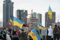 A crowd of bundled-up people stands outdoors in a city street during winter, waving multiple blue-and-yellow Ukrainian flags along with a Canadian flag. The gathering appears to be a rally or demonstration in support of Ukraine, with tall office buildings and streetlights in the background under an overcast sky.