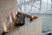 An elegant long table with people dining in an architecturally striking space, featuring tall glass windows and a high ceiling supported by metal beams.