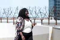 A smiling Black woman stands in front of a row of trees. She is looking to her left.