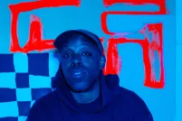 A blue-toned image of a black man wearing a cap and dark hoodie in front of a checkered backdrop with thick red lines.