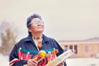 Elder Ma-Nee Chacaby stands outside surrounded by trees and cabin looking up at the sky playing a drum. 