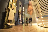 Five museum visitors look at towering exhibits in the “Indigenous Perspectives” gallery at the Canadian Museum for Human Rights. The nearest is made of wood and features trees, animals and a plaque. Behind the visitors, a rounded theatre built of bent wooden slats is visible.