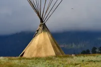 A large tipi sits in a field of grass. Its poles rise into a misty sky.