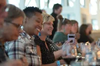 A group of people sit at a long dining table, laughing and enjoying a meal together. The focus is on two people smiling brightly - one wearing a plaid shirt and the other in a black patterned top, with glasses and plates arranged neatly in front of them.