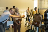 Students in a museum gallery display their work on easels. The focus is on one student talking with an adult who is smiling and taking a photograph with their phone of the student’s work.