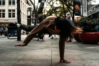 A woman in a grey jacket and darker grey pants is breakdancing on a sidewalk during daytime. Trees and buildings are in the background.