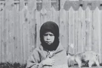 A young girl stands facing the camera with a sorrowful expression, clutching a shawl around her.