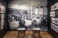 A museum exhibit that includes small school desks in front of a large photographic background of Indigenous children sitting at their desks in a classroom.