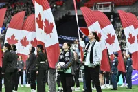People stand on a stadium field during a pre-game ceremony as large Canadian flags are held behind them, filling the background with red and white.