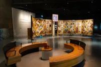 A museum exhibition featuring a large, curved freestanding wooden wall on which is mounted numerous small objects arranged in diamond and rectangular patterns. In the foreground, there circular benches creating an inviting space for visitors to view and contemplate the exhibit.