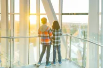 Two young children stand in a tower with their backs toward the camera as they stare out at a horizon overlooking a cityscape.