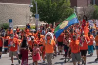 Hundreds of demonstrators in orange shirts, many also wearing traditional Indigenous articles such as a headdress or ribbon skirt and carrying signs, march down a wide paved street.