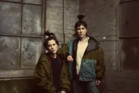 Two people with long hair in high ponytails wearing winter coats pose in the corner of a grey-painted brick room.
