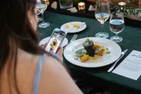 A woman with light skin and long, dark brown hair is using her cell phone to take a photo of a plate of gourmet food on a table.
