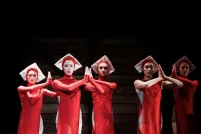 On a dark stage are five ballet dancers dressed in red knee-length flowy costumes with white square-like hats. They are looking forward with their hands in prayer.
