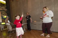 A young girl holds a small stick attached to a string and a small circular piece of cardboard. She is swinging this rope and cardboard as two adults watch. 