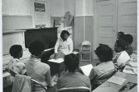 A black and white photo of a Black woman seated on a piano bench, twisted three-quarters towards a group of Black students. The students are seated in a semicircle in metal folding chairs and are holding notebooks. A table with two pens, a notebook with the top page filled with writing and a ripped piece of a poster is behind the students.
