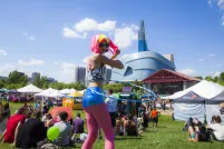 A hula hoop dancer with a pink and fluorescent yellow wig with pink leggings walks in a crowd of people sitting on a field of grass in front of a large glass building.