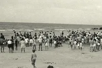 A black-and-white photograph of a crowd of people, most of them standing, on a beach.