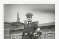 A black-and-white photo of a man in an army medic’s uniform featuring a white armband with a cross on it. Each side of the collar has a white patch with a large cross. The man is looking straight at the camera with a neutral expression. A building is behind him.