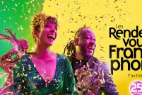 Two people dressed in chic clothes smile as confetti falls on them and hold theatre masks, in front of a colourful background.