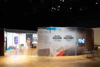 Upright panels are arranged in a circle in a dimly lit room. A gap offers people a way to enter the circle, and the interior of the circle is lit brightly. Next to the opening, a panel shows the words "Behind Racism: Challenging the Way We Think."