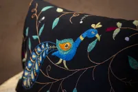 Close-up photo of a rectangular black throw pillow resting on a wooden bench, featuring detailed embroidery of a stylized peacock with a curved blue neck, teal head crest, and an orange-and-teal eye-patterned feather. The design also includes brown vine-like stitching, teal and sage leaves, and small maroon and white floral accents scattered across the black fabric.