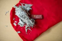 A small bundle of dried sage lies on a bright red cloth, with loose leaf fragments scattered around. Beside it is a small rectangular match box labeled “Mother Earth Tobacco.“ The items rest on a light wooden surface.
