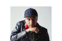 Tom Morello looks intently at the camera while leaning on his hand, which holds a guitar by the neck. He is wearing a black leather jacket, red collared shirt and baseball hat.