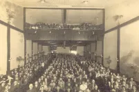 A black and white photo of a movie theatre audience. The picture is taken from the front of the theatre looking towards the back, so the faces of the audience can be seen. Potted palm trees line the walls on each side.