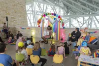 A drag artist reads aloud to a group of children and adults seated in colourful chairs inside a bright, industrial-style venue decorated with rainbow banners and bunting fans for a Pride-themed storytime event.