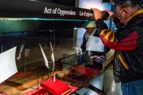 A man with long, braided hair bows his head in front of a glass-walled display where two feathers are lodged.
