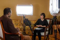 A man and a woman sit in chairs facing each other. There is a microphone on a stand positioned between them. There are also large lights on both sides of the woman’s chair, which are pointed towards the man.