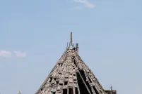 A large, weathered wooden structure shaped like a tepee stands in a dry grassy field under a clear blue sky. The structure's wooden frame is largely exposed, with many wood shingles missing or falling off, revealing the ribbed skeleton beneath.