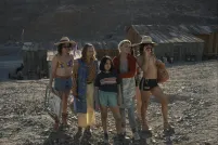 Five members of a queer family stand together on a rocky hillside in front of weathered wooden shanties in a desert mining town, with a young girl at the centre of the group looking in the same direction just off-camera.