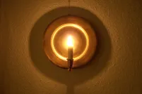 Lit candle on a round brass wall sconce, its reflector glowing gold and casting a circular shadow on a textured wall.
