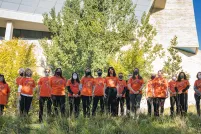 People wearing orange T-shirts stand in front of a row of trees. There is long grass in the foreground. The T-shirts bear images of small hand prints grouped in a heart shape with the number 215+ above them.