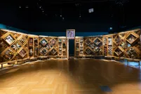 A large, curved freestanding wooden wall on which is mounted numerous small objects arranged in diamond and rectangular patterns. There is an open door in the centre of the wall.