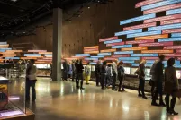 A large crowd in a Museum gallery. Most people are looking at a display on the wall of the gallery. The display consists of a series of pictures and of long rectangular text panels placed horizontally.