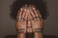 Aaju Peter covers her eyes and cheeks with her bare hands. Tattoos are visible on her forehead, chin, fingers and wrists. Her hands and the visible parts of her face are illuminated and are framed by her dark curly hair and angular shoulders.