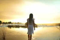 A girl with long dark hair wearing a knee-length white dress faces away, standing ankle deep in a calm lake, with the setting sun casting a warm yellow glow across the water.