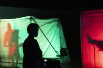 The silhouette of a person standing in the dark in front of red and green clothed box structures lit from the inside and containing shadowed figures.