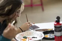 A young woman sitting at a table paints a design on a tile while a tin tray with various paints sits in front of her. An open bottle of red paint sits on the table. 
