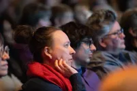Audience members sit in a dimly lit venue watching a presentation, their faces illuminated by stage light. In the foreground, a person with their hair in a bun and wearing a bright red scarf rests their chin on their hand, looking attentively toward the stage. Other attendees sit beside them in soft focus, creating a quiet, contemplative atmosphere as the crowd listens.